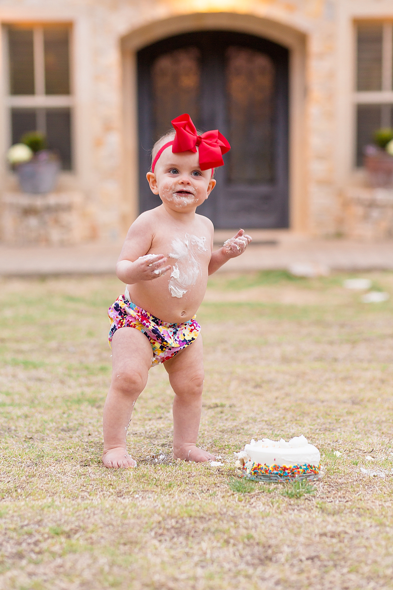 Toddler in colorful bloomers and bow has first birthday cake smash in Adriatica Village with McKinney family photographer, Wisp + Willow Photography Co. 