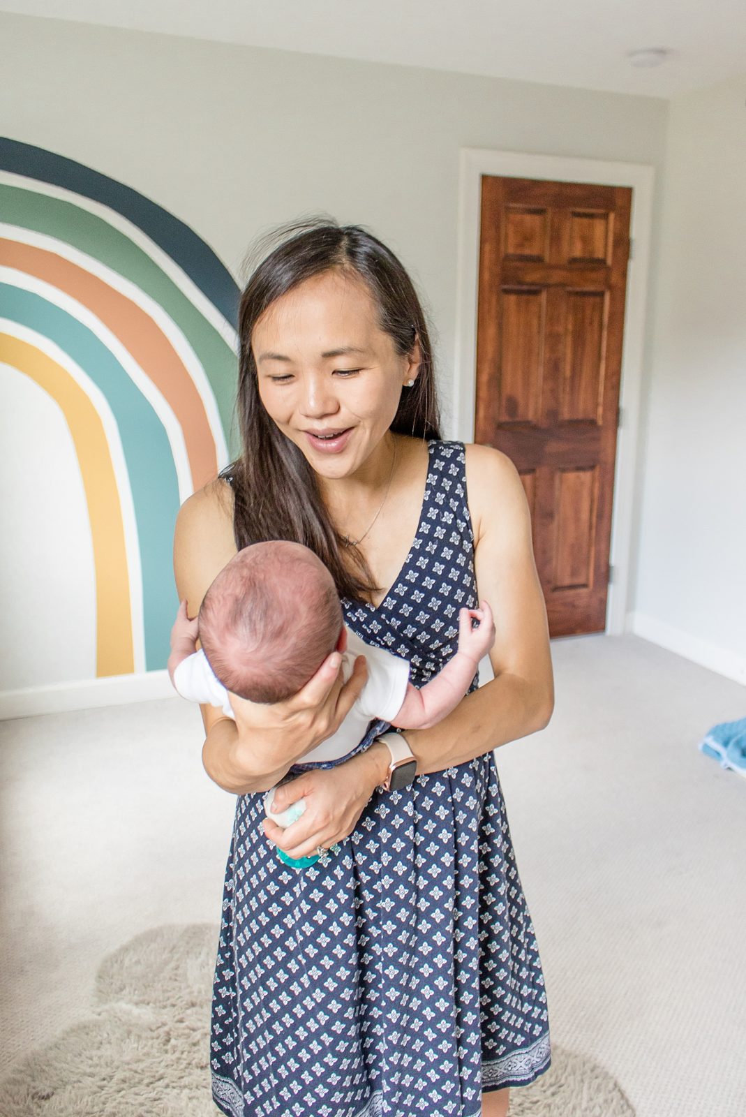 mom holds newborn son in nursery with rainbow on wall during lifestyle session with Brentwood family photographer Wisp + Willow Photography Co.