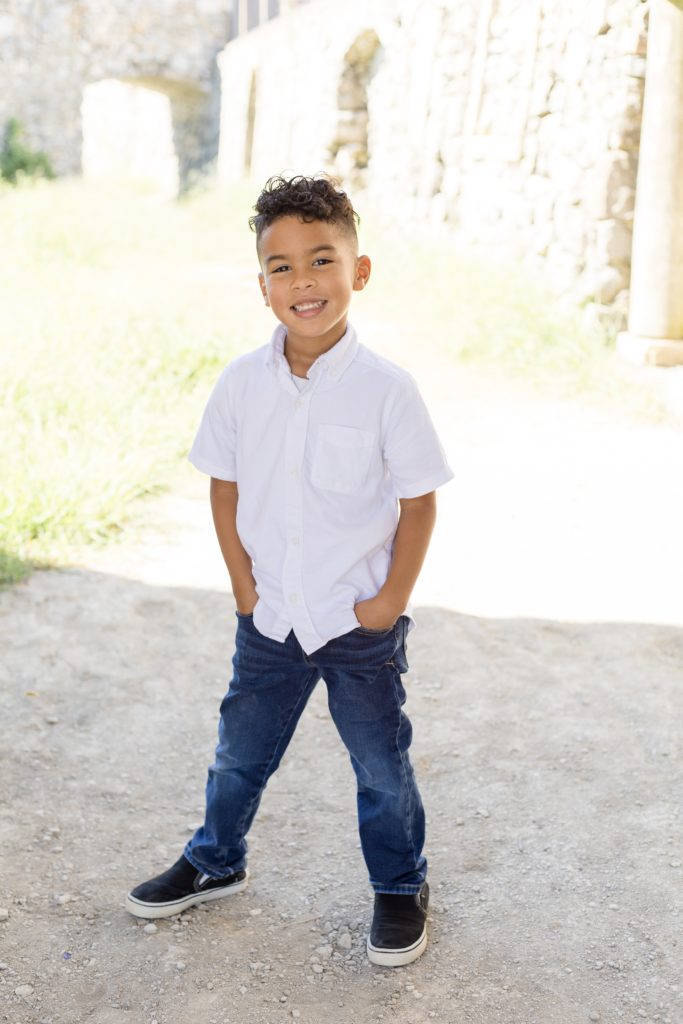 Young boy in short sleeve button up shirt and jeans poses alone during fall family portrait session at Adriatica Village in McKinney, TX with McKinney maternity photographer Wisp + Willow Photography Co.