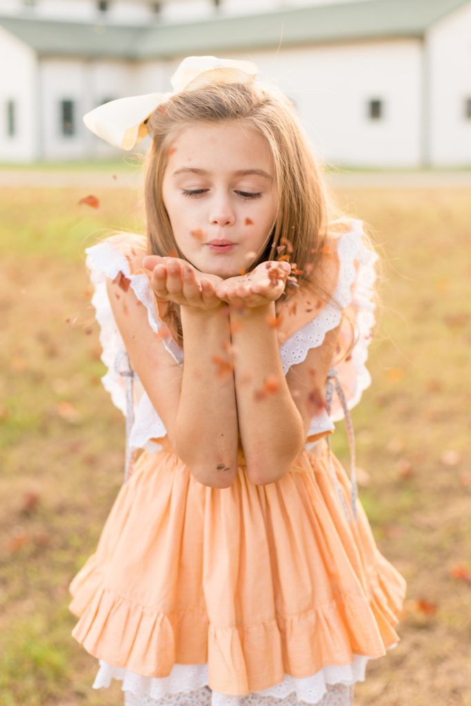 Girl in orange dress blows leaves out of her hand for creative photo during family portrait session in the fall at Harlinsdale Farm in Franklin, TN. Click to see more from this family session shot by Wisp + Willow Photography Co.