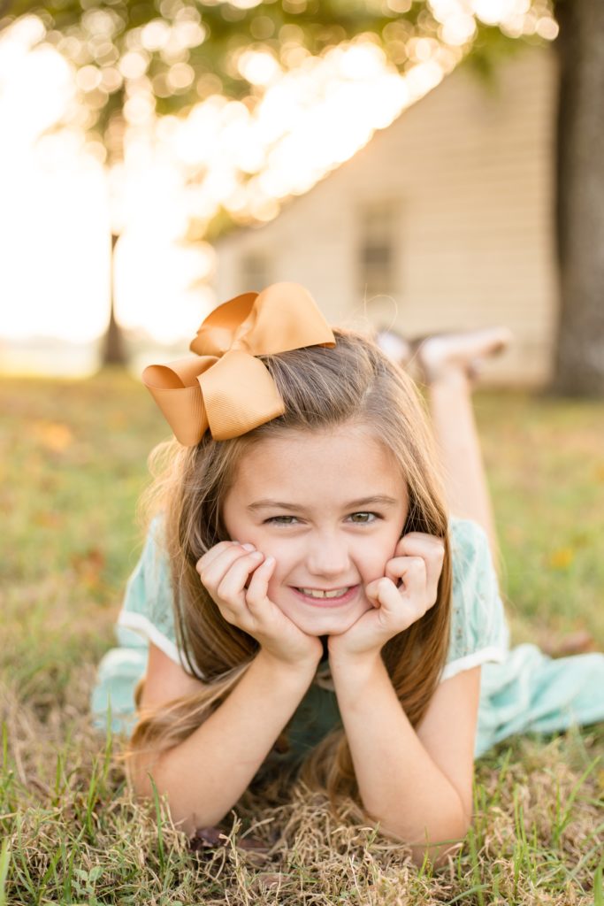 Girl in green dress and peach bow lays in grass in front of farm house at Harlinsdale Farm in Franklin, TN for family portrait session. Click to see more from this family portrait session shot by associate photography team Wisp + Willow Photography Co.