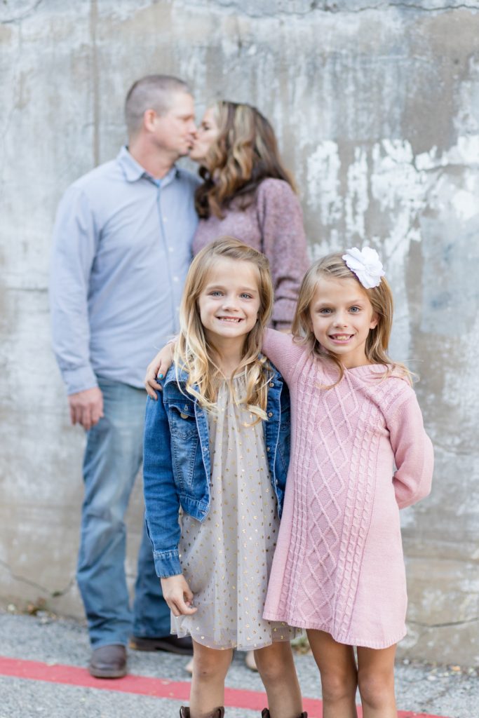 Mom and dad kiss in the background while sisters pose for photo in historic Downtown McKinney Square for a family portrait session with Wisp + Willow Photography Co.