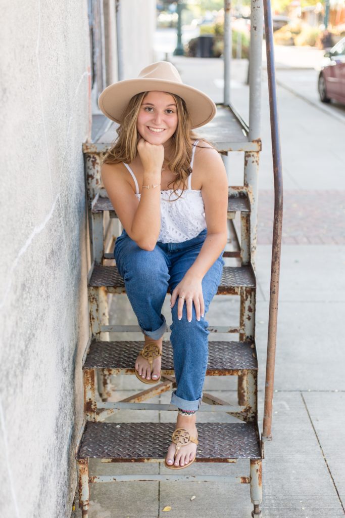 Senior poses on stairs in Historic Downtown McKinney Square for senior portrait session with Wisp + Willow Photogrpahy Co. She wore a white cropped shirt with jeans and tan sandals.