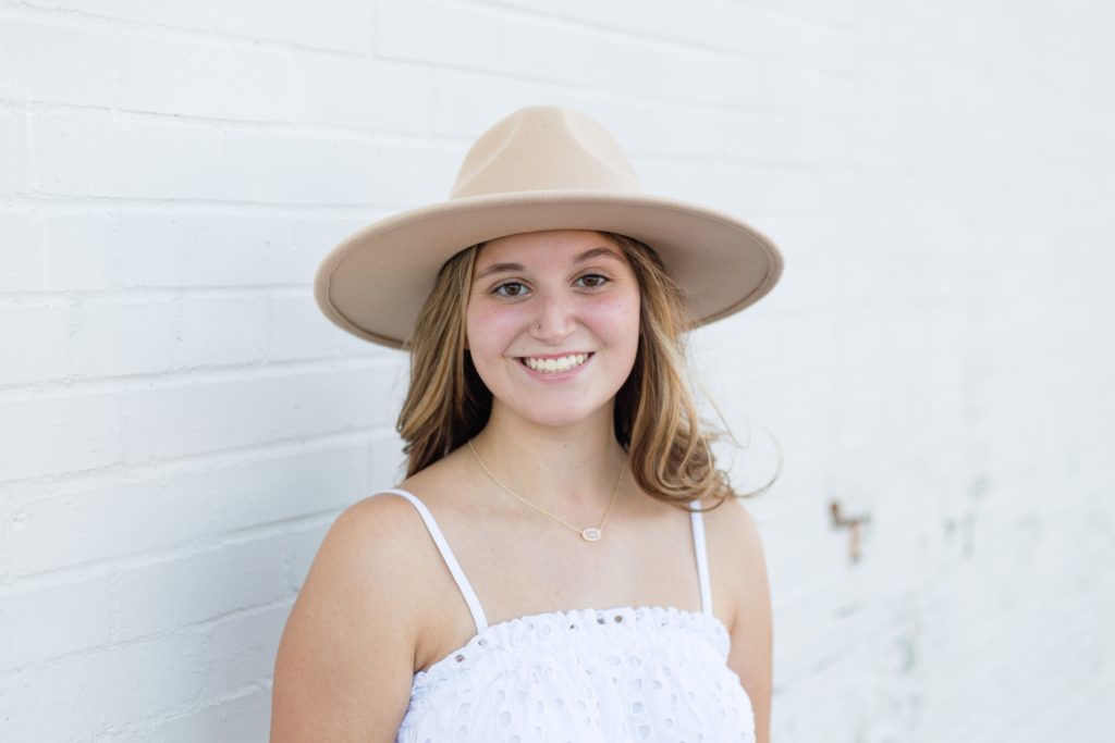 Senior poses in front of white wall in tan hat and white tank top for senior portrait session with Wisp + Willow Photography Co. in the historic Downtown McKinney Square. Click to see more from this senior session!