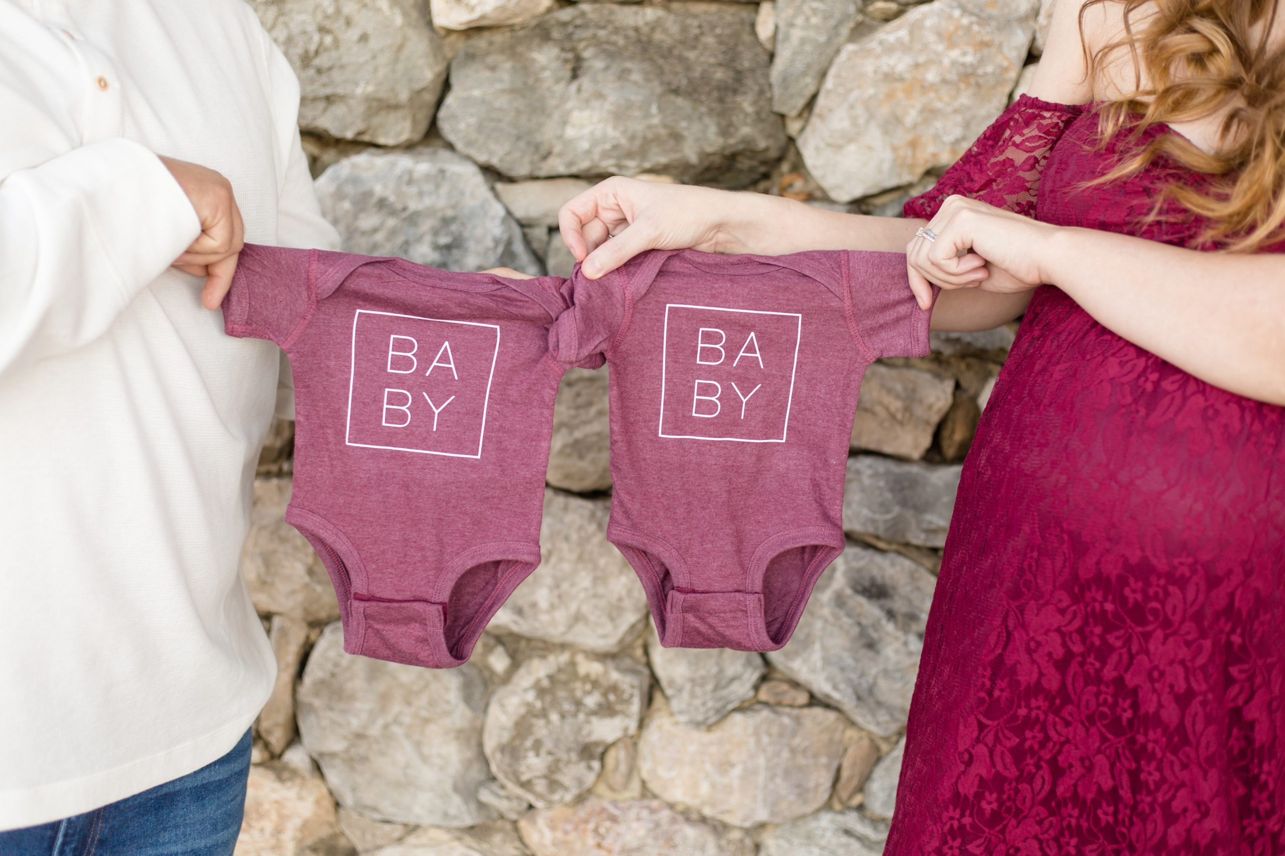 Expecting parents hold maroon onesies during maternity session with Wisp + Willow Photography Co. at Adriatica Village in McKinney, TX.