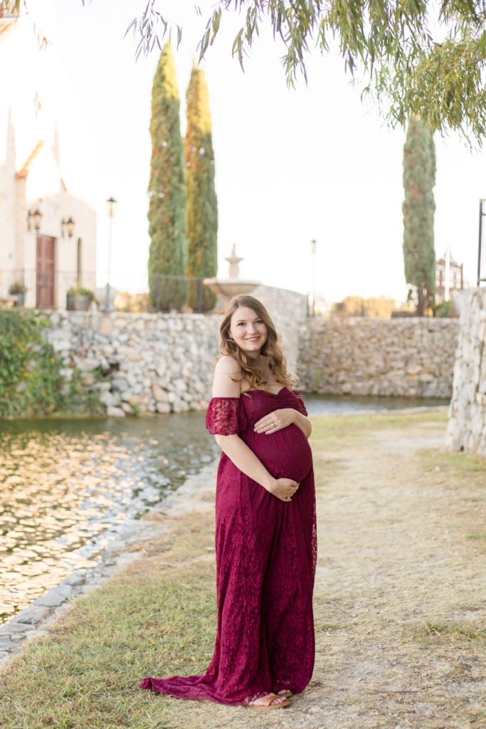 Expecting mom in maroon lace gown holds belly in front of Bella Donna Chapel at Adriatica Village in McKinney, TX during maternity session with Wisp + Willow Photography Co.