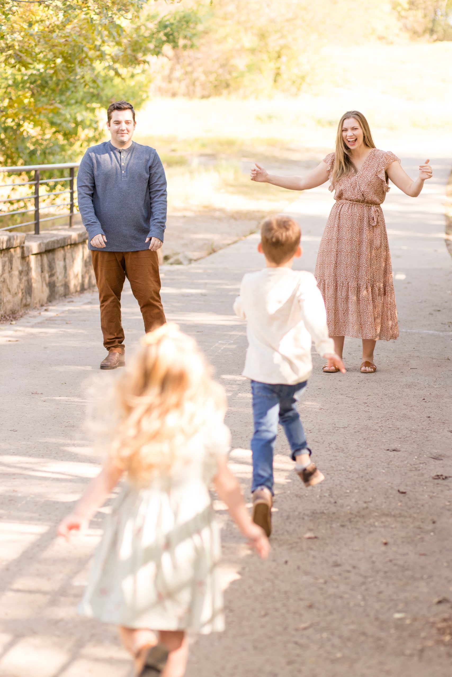 Parents stand while kids run to them for candid shot during family photo session in the fall at Arbor Hills Nature Preserve