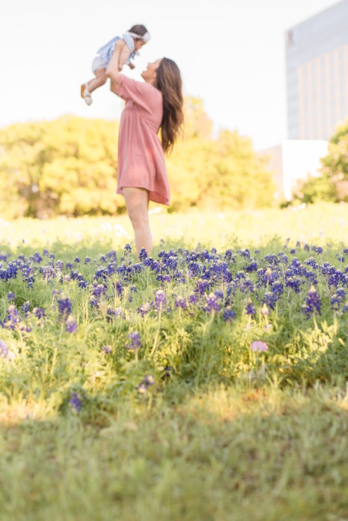 mom in pink dress holds daughter in the air in field of bluebonnets in Plano, TX during bluebonnet mommy & me session. Click to see more from this session on our blog!