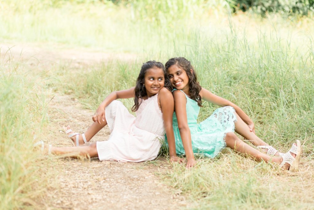 Sisters sit on ground back to back and look at each other during spring family portrait session at Arbor Hills Nature Preserve in Plano, TX with Wisp + Willow Photography Co. Click to see more from this sweet session live on our blog now! 