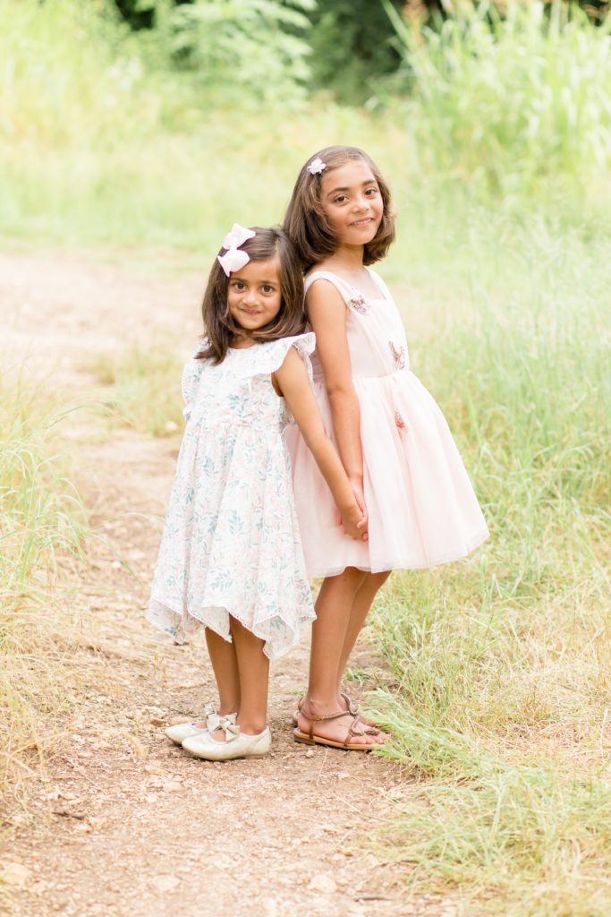Little girls in dresses hold hands and stand back to back during family portrait session in the spring at Arbor Hills Nature Preserve in Plano, TX with family photography team Wisp + Willow Photography Co. Click to see more from this sweet session on our blog now! 