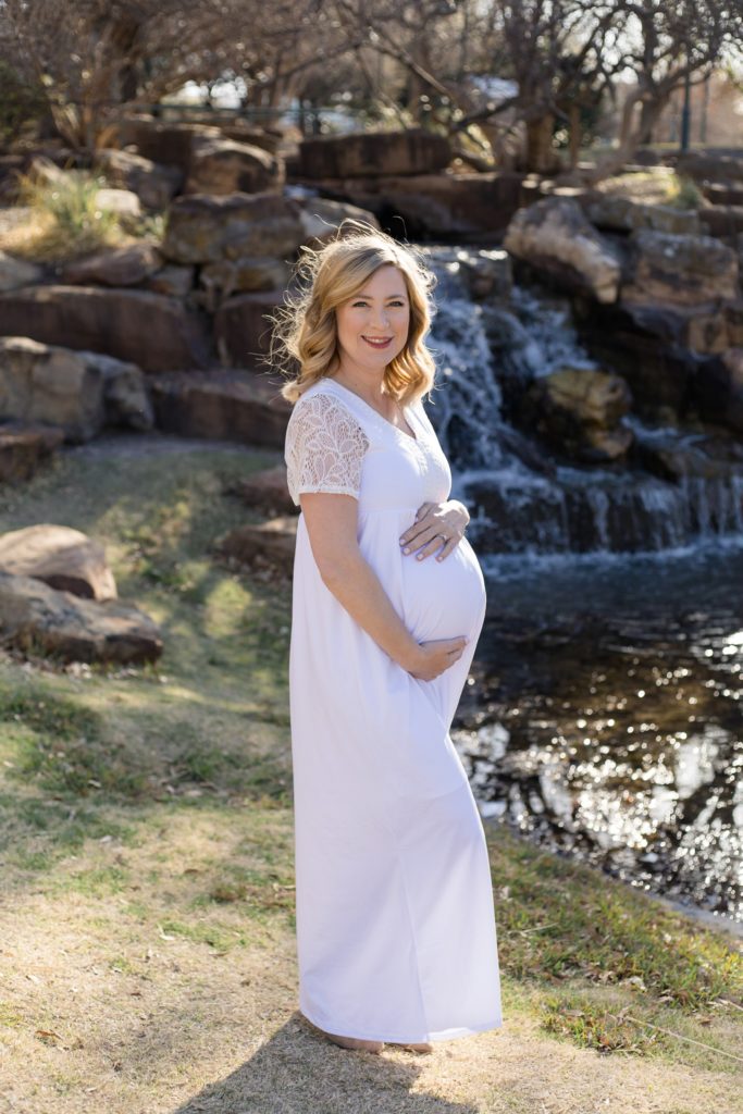 Mom holds pregnant belly in white dress in front of waterfall at Frisco Central Park in Frisco, TX during spring maternity session with family photography team Wisp + Willow Photography Co. 