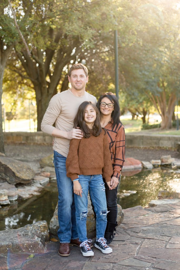 family of 3 poses in front of stream at Frisco Central Park in Frisco, TX during fall minis with family photography team Wisp + Willow Photography Co.