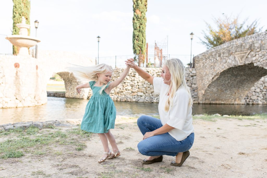 Mom holds daughter's had while she twirls around in green dress in front of Bella Donna Chapel at Adriatica Village in McKinney, TX. Click to see more from this sweet family portrait session on the blog now!