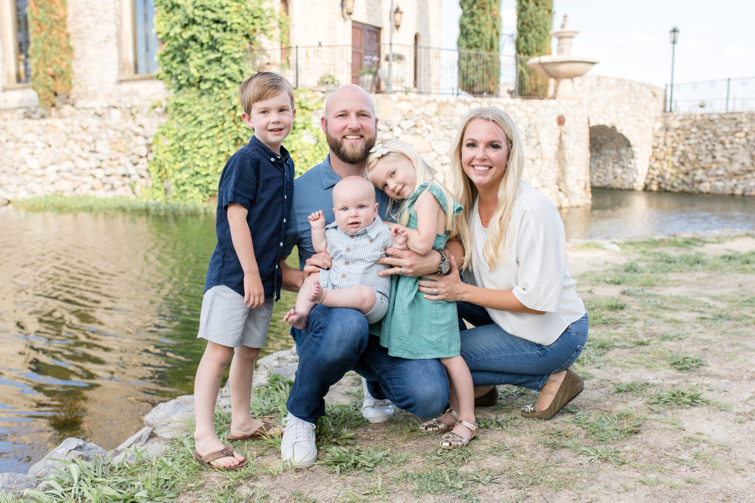 Family of 5 poses on the ground in front of Bella Donna Chapel next to the reservoir at Adriatica Village in McKinney, TX. Click to read more about this sweet session live on the blog now.