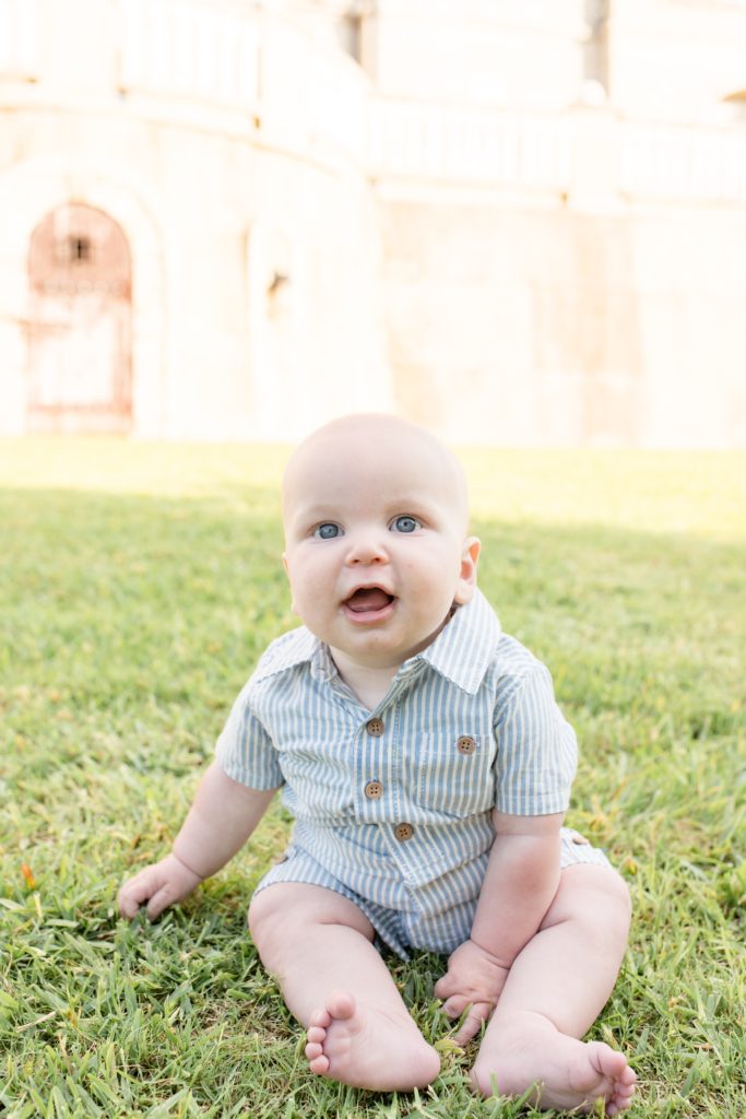Baby boy sits in grass in front of building at Adriatica Village during family portrait session with family photography team Wisp + Willow Photography Co. Click to see more from this sweet session live on the blog now!