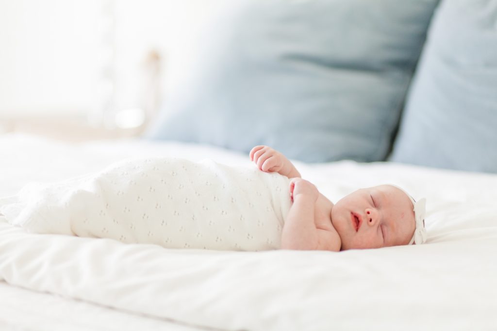 Sleeping newborn in eyelet swaddle and white bow lays on parent's bed during lifestyle newborn session in Raleigh, NC in the spring with family photography team Wisp + Willow Photography Co. Click to read more about this precious session photographed by our associate team on our blog now! 