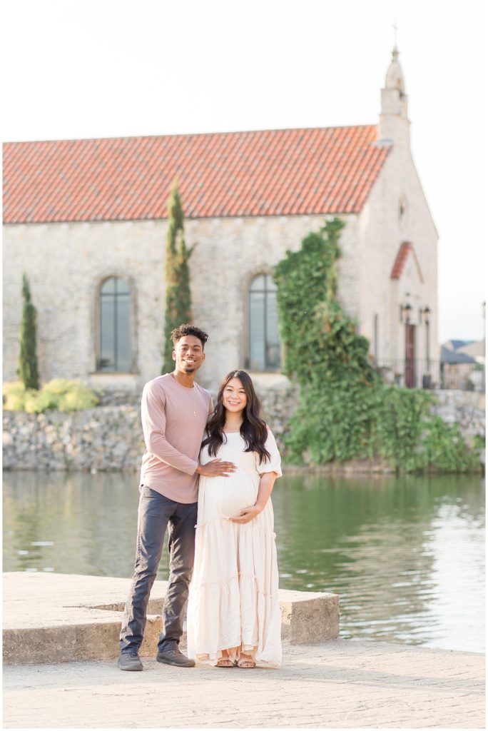  Adriatica Village | Wisp + Willow Photography Co. Beautiful couple standing in front of pond at sunrise. Mom to be is wearing a long cream dress. Dad to be is dressed in a dusty rose long sleeve top.