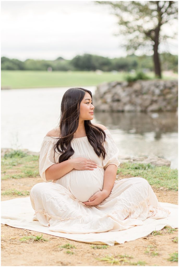  Adriatica Village| McKinney, Tx | Wisp + Willow Photography Co. Mom is seated on the ground in front of pond. She is wearing a beautiful off the shoulder cream dress.