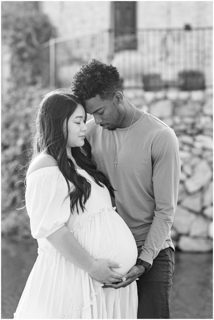 Adriatica Village | McKinney, TX | Wisp + Willow Photography Co. Couple standing with heads together and eyes closed with stone bridge in the background and holding baby bump.