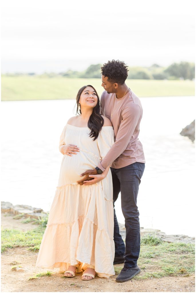 Adriatica Village | McKinney, TX | Wisp + Willow Photography Co. precious couple standing outside looking at each other and holding baby bump. Mom is wearing long cream off-the-shoulder dress and dad is wearing dusty rose long-sleeved shirt and blue jeans. 