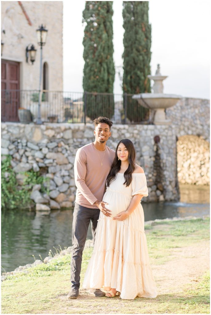 Couple is standing outside in front of pond and stone bridge in McKinney, TX Mom and dad are sweetly holding baby bump. Mom is dressed in long cream off-the-shoulder dress and dad is wearing long-sleeved dusty rose shirt. Maternity session with Wisp + Willow Photography Co.