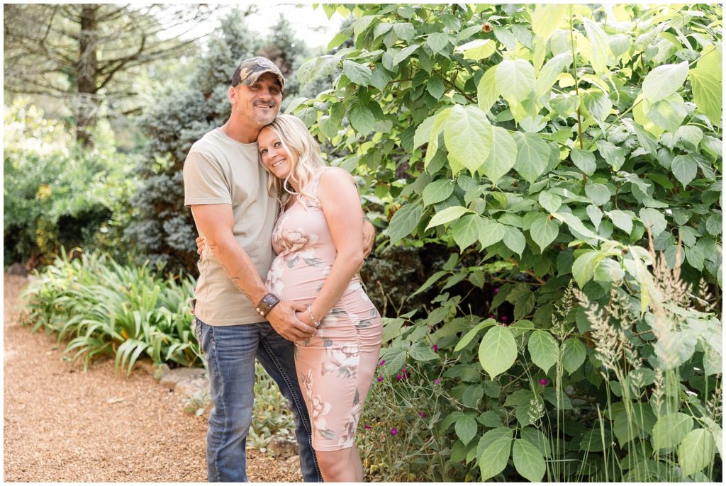 Parents to be are standing on a beautiful trail surrounded by lush greenery at Botanical Gardens| Knoxville, TN | Wisp + Willow Photography Co. Mom is wearing a beautiful blush pink floral dress and sandals. Dad is wearing a sage shirt and blue jeans.