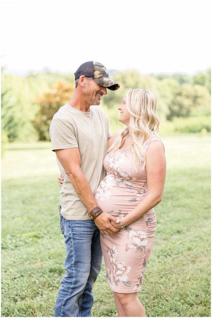 Mom and dad are pictured in an open field at Botanical Gardens Maternity Session| Knoxville, TN | Wisp + Willow Photography Co. Mom is wearing a blush pink floral sleeveless dress. Dad is wearing a sage shirt, blue jeans, and a cap.