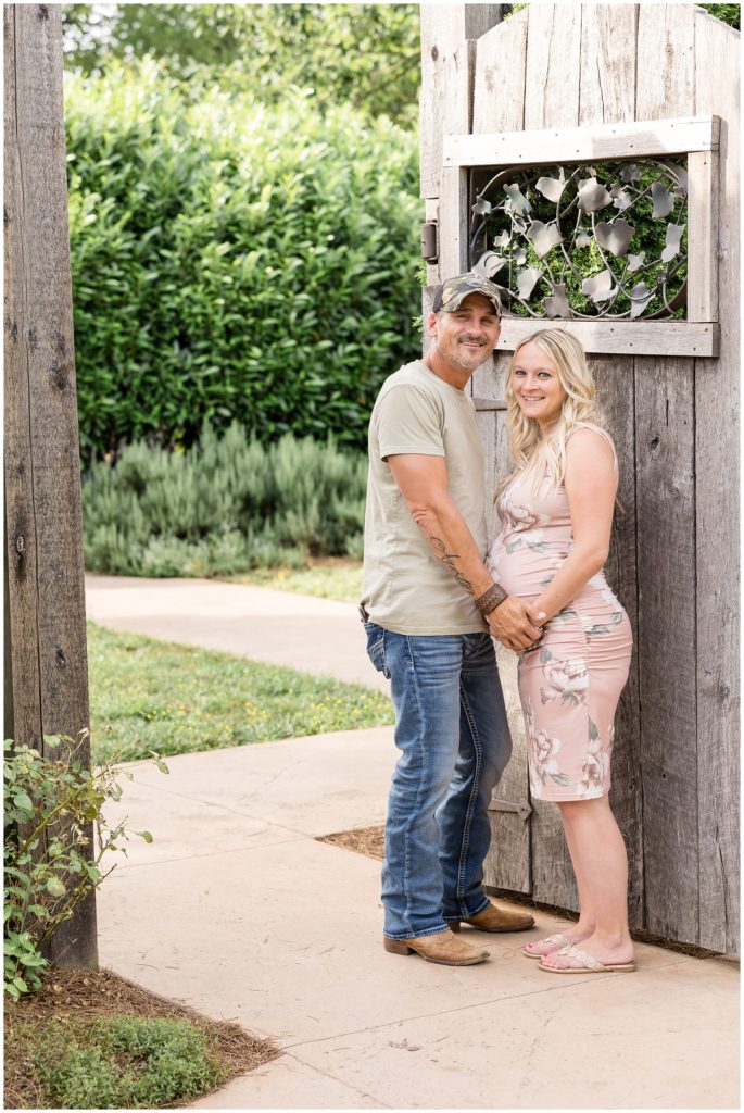 Mom and dad are standing in front of a wooden gate with intricate iron leaves at Botanical Gardens Maternity Session| Knoxville, TN | Wisp + Willow Photography Co. Mom is wearing a beautiful blush floral pink sleeveless dress and white sandals. Dad is wearing a sage shirt, blue jeans, brown boots, and a cap.