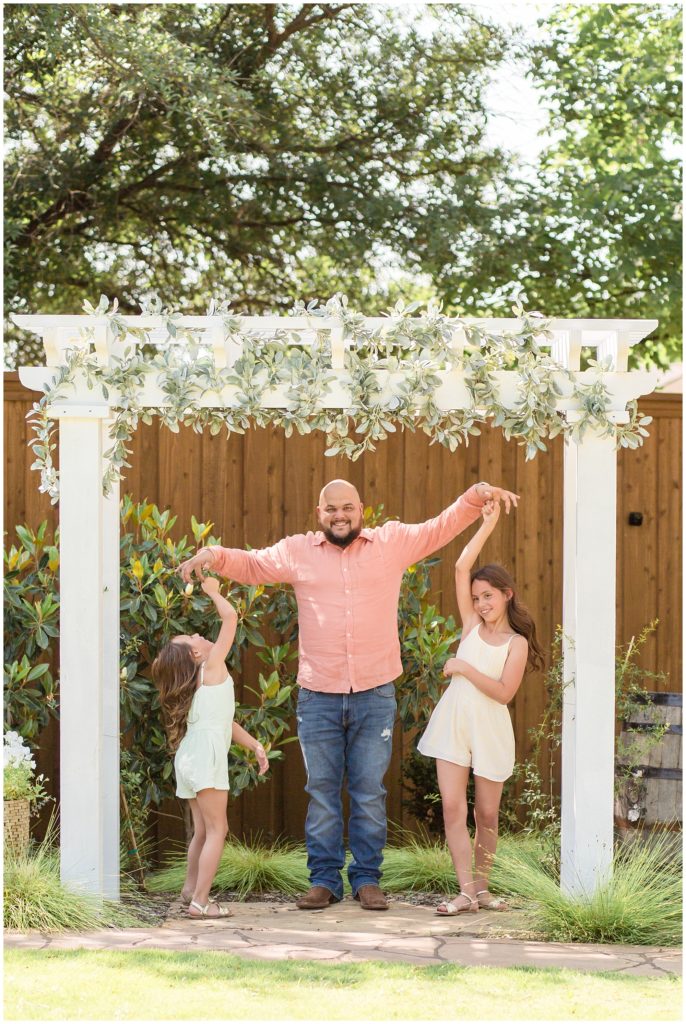 Dad and daughters are seen dancing below white columns with hanging vines. There is a rustic wooden fence and wooden barrel in the background. The first sister is wearing a light green romper and gold sandals. Dad is wearing a pink long-sleeved button-up shirt with blue jeans and brown boots. The second sister is wearing a cream-colored romper and gold sandals.