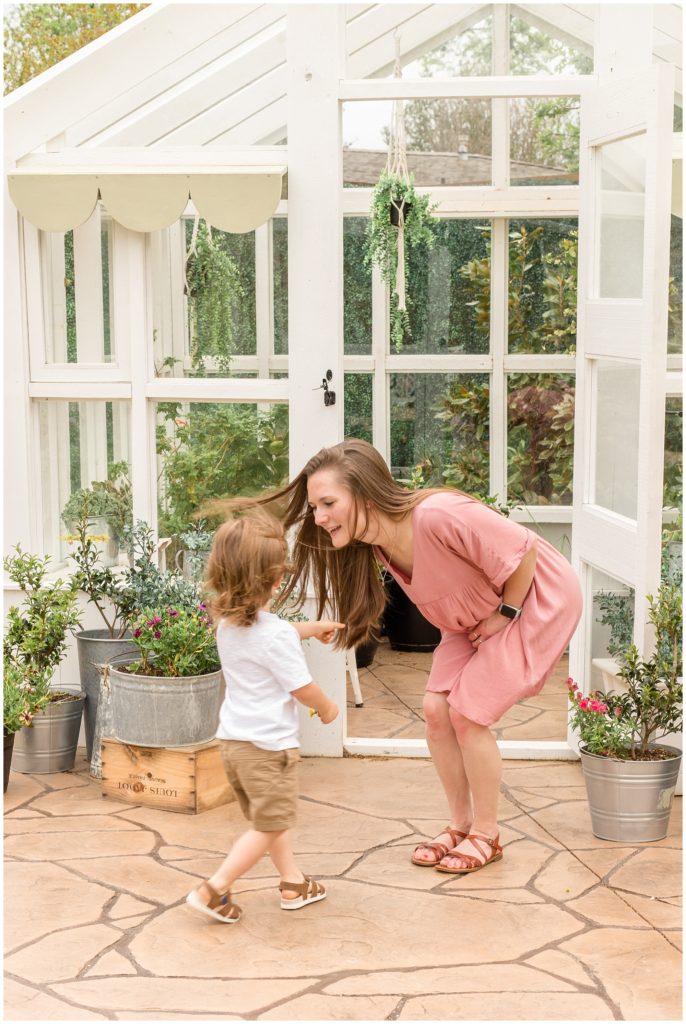 Adorable mom and son are seen playing just outside a gazebo decorated with beautiful greenery. Mom is wearing a light pink short-sleeved dress and brown sandals. Son is wearing a short-sleeved white shirt, khaki shorts, and brown sandals.