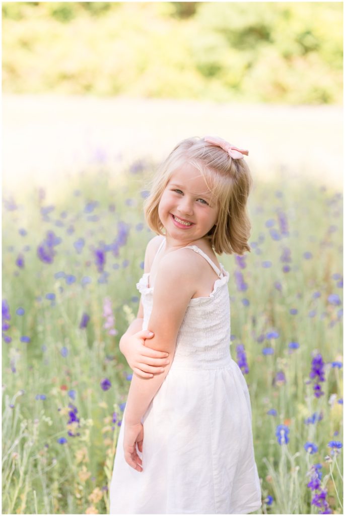 Blonde girl with a pink bow in a white sleeveless dress with straps looking over her shoulder and standing in wildflowers during her Texas Wildflower Mini in Richardson, TX with Wisp + Willow Photography Co.