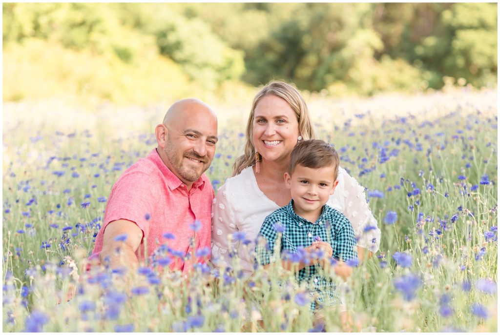 sweet family of three dad in pink button up collared shirt mom in white dress with sheer sleeves and white flowers little boy with blue and green checkered button up shirt sitting in the wildflowers during their Texas Wildflower Mini in Richardson, TX with Wisp + Willow Photography Co.