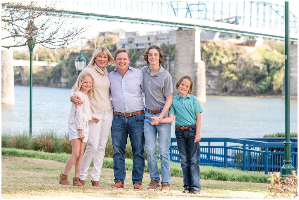 A beautiful family of 5 is seen here standing with a large bridge and lake in the background. The daughter is wearing a light cream-colored long-sleeved shirt, white denim short skirt, and brown booties. Mom is wearing a cream-colored long-sleeved shirt with a cowl collar, white pants, and brown booties. Dad is wearing a long-sleeved button-up light blue shirt, blue jeans, and brown boots. The first son is dressed in a grey polo shirt, blue jeans, and brown shoes. The second son is wearing a green polo shirt, blue jeans, and black shoes. Coolidge Park Family Session | Chattanooga, TN