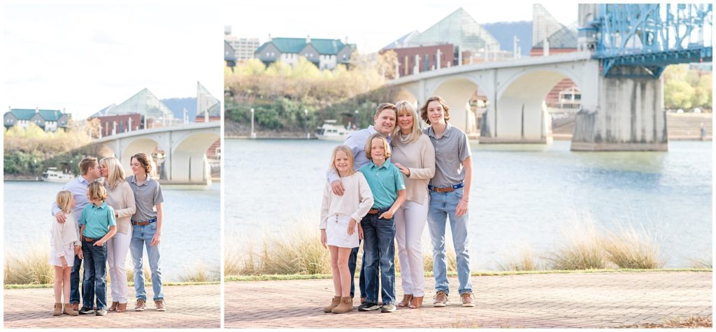 This amazing family of 5 is pictured here with a large bridge, lake, and adorable houses just in the background. The daughter is wearing a light cream-colored long-sleeved shirt, white denim short skirt, and brown booties. Mom is wearing a cream-colored long-sleeved shirt with a cowl collar, white pants, and brown booties. Dad is wearing a long-sleeved button-up light blue shirt, blue jeans, and brown boots. The first son is dressed in a grey polo shirt, blue jeans, and brown shoes. The second son is wearing a green polo shirt, blue jeans, and black shoes. Coolidge Park Family Session | Chattanooga, TN