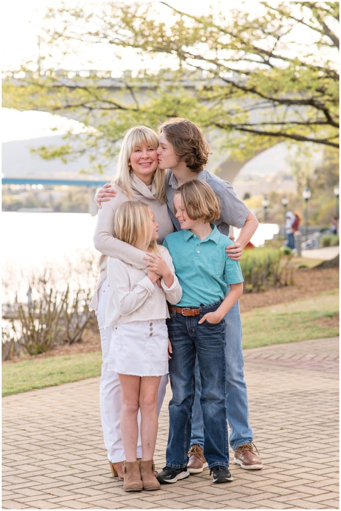 Mom and her kids are standing embraced with the lake, bridge and trees just in the background. The daughter is wearing a light cream-colored long-sleeved shirt, white denim short skirt, and brown booties. Mom is wearing a cream-colored long-sleeved shirt with a cowl collar, white pants, and brown booties.  The first son is dressed in a grey polo shirt, blue jeans, and brown shoes. The second son is wearing a green polo shirt, blue jeans, and black shoes. Coolidge Park Family Session | Chattanooga, TN