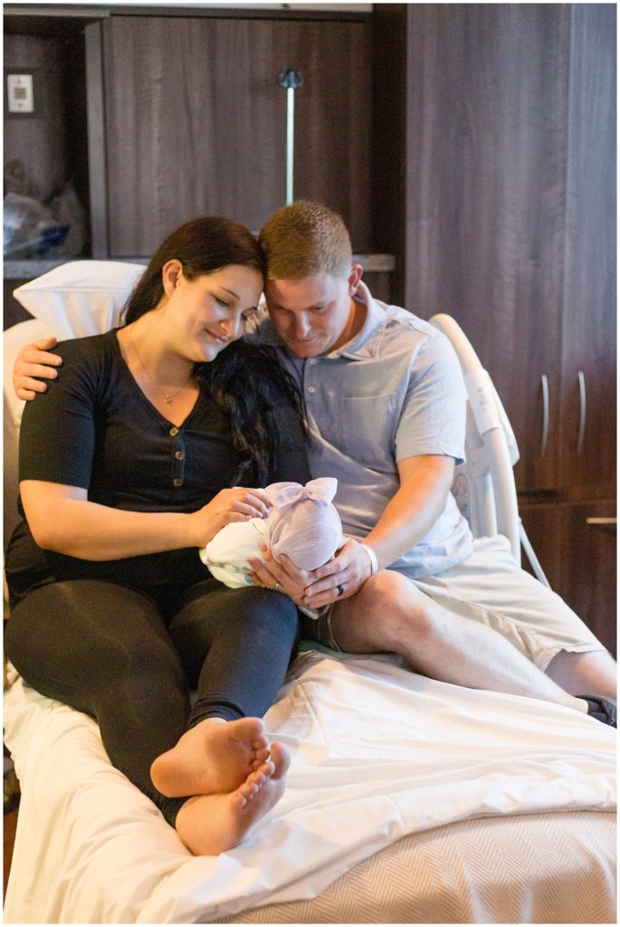 Precious family of 3 in their Fresh 48 session. Mom is dressed in a short-sleeved black shirt with brown buttons and black leggings. Dad is wearing a light blue polo shirt and khaki shorts. The baby girl is wearing a pink cap with a matching bow and is swaddled in a white blanket.