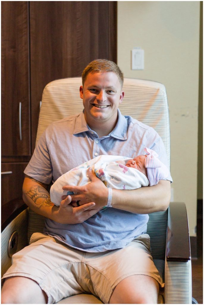 Dad is so proud holding his little girl! Dad is wearing a short sleeve light blue polo with khaki shorts. Baby girl is wearing a pink hat with a matching bow and is swaddled in a white blanket.