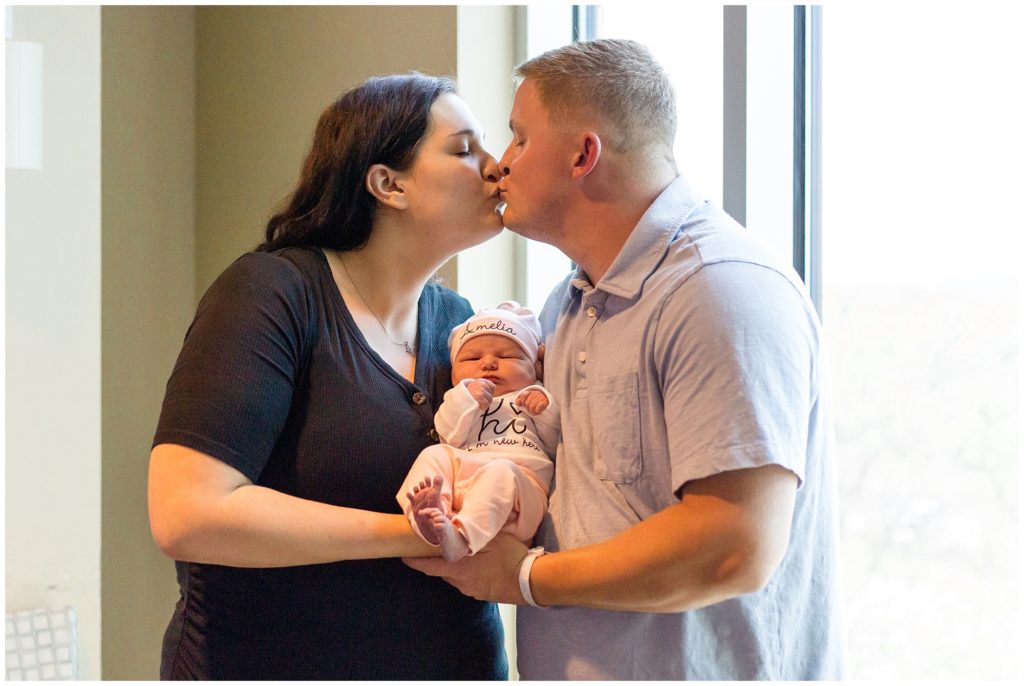 Mom and dad share a kiss while holding sweet baby girl! Mom is wearing short sleeve black shirt. Dad is wearing short sleeve light blue polo. Baby girl is wearing a pink cap, white onesie, and pink pants.