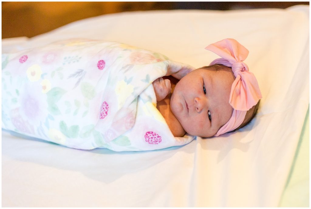 Sweet baby girl is laying on a white blanket. She is swaddled in a white blanket with a pink floral print and a pink bow headband.