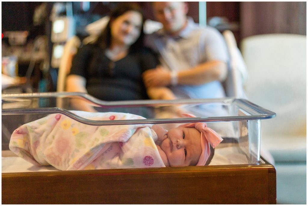 Sweet baby girl is the focus of the precious photo of the new family of 3. Baby is swaddled in a blanket with a floral print and pink bow headband. Mom is wearing a black short-sleeved shirt. And dad is wearing short-sleeve blue polo.