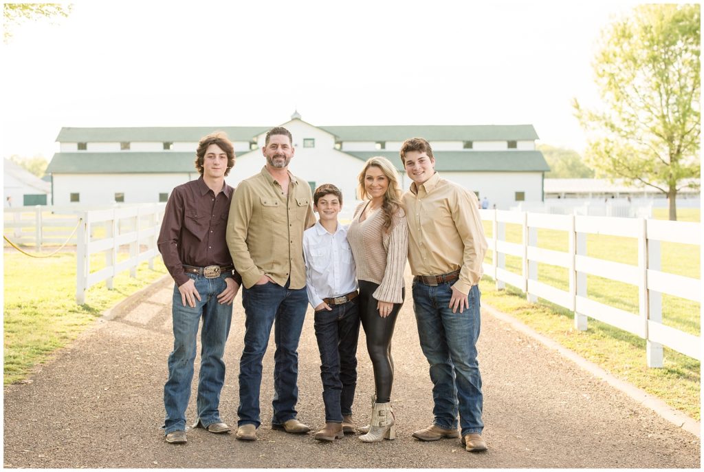 A beautiful family of 5 is standing on a gravel road between a white wooden fence and an amazing white barn in the background. The first son is wearing a dark brown long-sleeved button-up shirt with blue jeans and brown boots. Dad is wearing a long-sleeved light brown button-up long-sleeved shirt with blue jeans and brown boots. The next son is wearing a long-sleeved button-up white shirt with blue jeans and brown boots. Mom is dressed in a long-sleeved cream-colored sweater with black leggings and cream-colored buckle booties. The last son is wearing a light brown long-sleeved button-up shirt and blue jeans with brown boots.