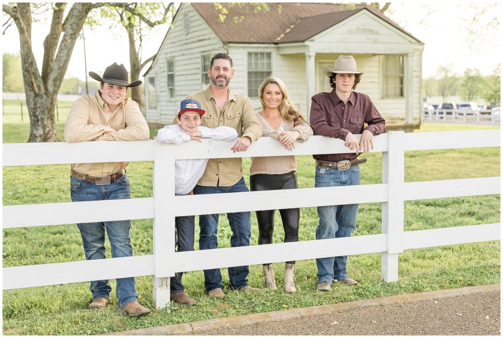 A family of five is pictured leaning on a white wooden fence with a rustic white wooden house in the background at Harlinsdale Farm | Franklin, TN. The first son is wearing a light brown long-sleeved button-up shirt with blue jeans, brown boots American flag belt buckle, and a dark brown cowboy hat. The next son is wearing a long-sleeved button-up white shirt with blue jeans and brown boots ad a red, white, and blue cap. Dad is wearing a long-sleeved button-up light brown shirt, blue jeans, and brown boots. Mom is wearing a cream-colored sweater black leggings and cream-colored booties. The last son is wearing a dark brown button-up long-sleeved shirt, blue jeans, brown boots, and a light brown cowboy hat.
