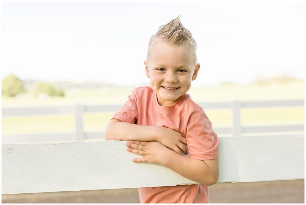 Adorable little boy is pictured at Harlinsdale Farm | Franklin, TN hanging on a white wooden fence. This precious boy is wearing a salmon-colored short-sleeved t-shirt and the sweetest smile!
