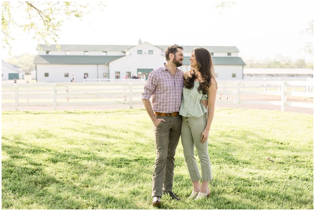 A lovely couple is seen here at Harlinsdale Farm | Franklin, TN. They are standing in a field just in front of a wooden fence and a gorgeous white barn in the background. The husband is wearing a white and purple checkered button-up shirt with the sleeves rolled up and olive green pants. The wife is wearing a sleeveless white shirt with sage green details and sage green pants and cream-colored slides.