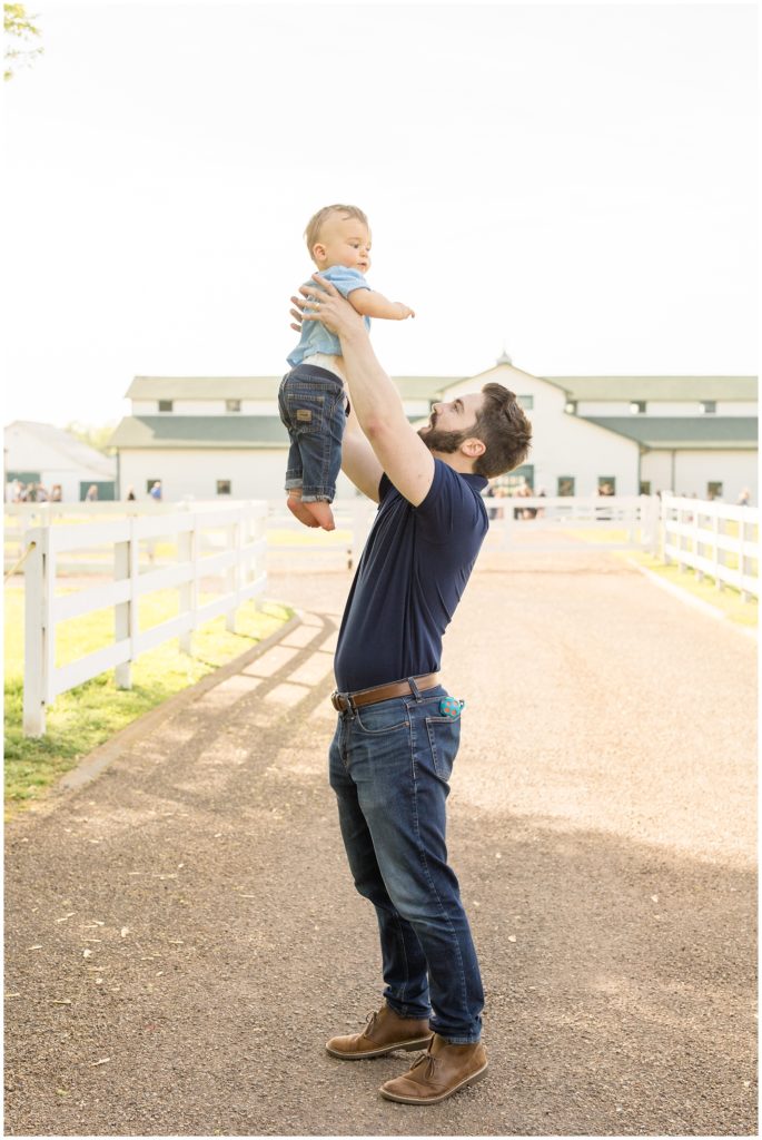 Father is playing with son here at  Harlinsdale Farm | Franklin, TN. There is a beautiful white barn in the background and standing in between white wooden fence. Dad is wearing short-sleeved navy polo, blue jeans and brown shoes. Baby is wearing light blue shirt and blue jeans.