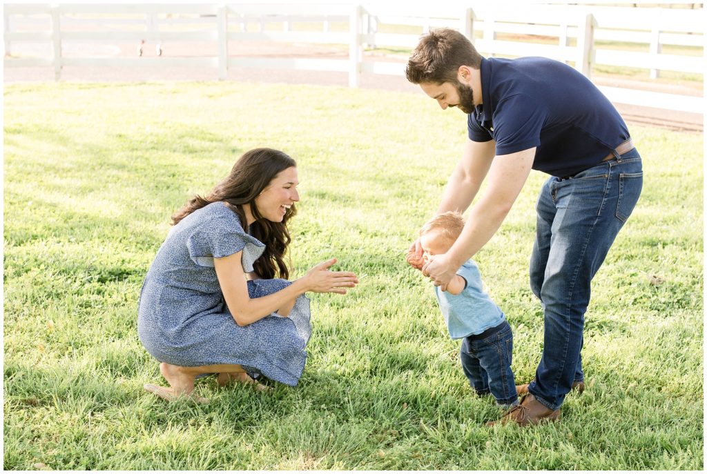 Family of 3 playing in a field with a white wooden fence in the background. Mom is kneeling and clapping as her son walks toward her. She is wearing a blue dress with ruffle cap sleeves. Son is wearing a light blue polo and blue jeans. Dad is holding their son's hands and helping him walk to mom. Dad is wearing a navy polo shirt and blue jeans with brown shoes. 