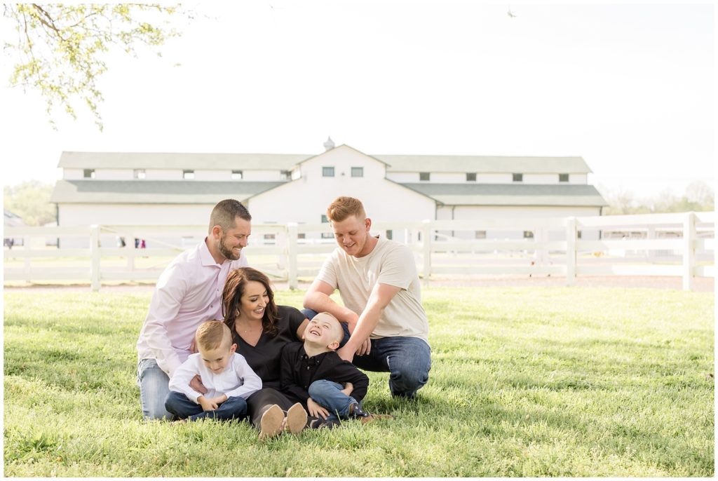 A beautiful family of 5 pictured sitting and playing in a field with a white wooden fence and a gorgeous white wooden barn in the background. Dad is wearing a long-sleeved button-up shirt and blue jeans. Mom is wearing a long black satin dress. The first brother is wearing a white button-up long-sleeved shirt and blue jeans. The second brother is wearing a long-sleeved button-up black shirt and blue jeans. The last brother is wearing off white short-sleeved shirt and blue jeans. 