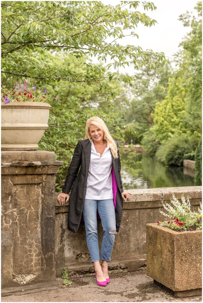 The client is seen here leaning against a beautiful stone bridge with oversized planters and huge hanging trees. She is wearing a white shirt with a collar, blue jeans, a long black jacket lined in hot pink, and matching hot pink high heels.