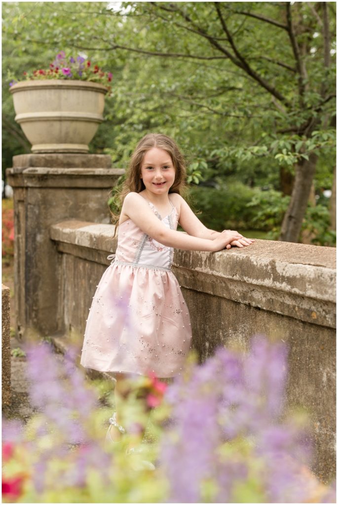 This precious little girl is standing on a stone bridge with an oversized plantar and huge trees in the background. She is wearing a beautiful soft pink sleeveless dress with tiny pearl embellishments. Centennial Park | Nashville, TN 