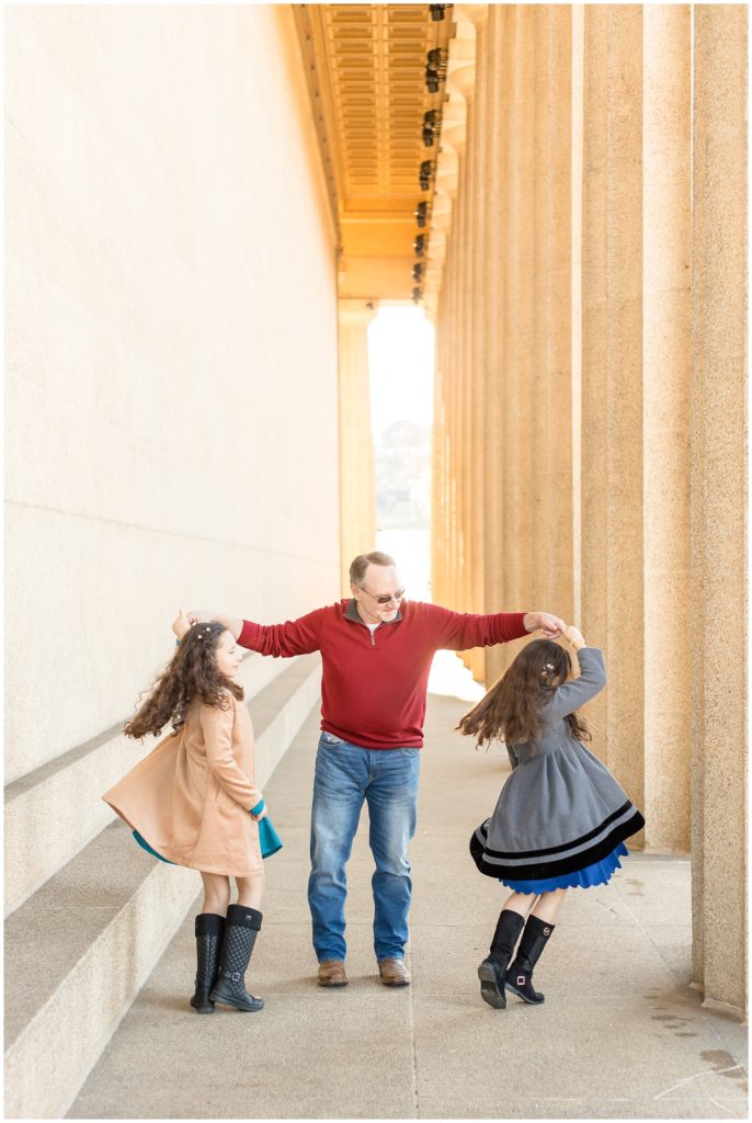 A dad and his daughters are dancing in this amazing archway. The first daughter is wearing a tan peacoat with a teal dress underneath and black boots. Dad is wearing a red long-sleeved pullover shirt, blue jeans, and boots. The second daughter is wearing a grey peacoat with black trim, a royal blue dress, and black boots.